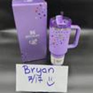 Puede incluir: Un vaso t&eacute;rmico morado con asa, tapa abatible y base blanca, junto a una caja morada a juego. El vaso y la caja presentan un dise&ntilde;o moteado. Una nota manuscrita con el nombre "Bryan" y la fecha 7/17 est&aacute; en la imagen.