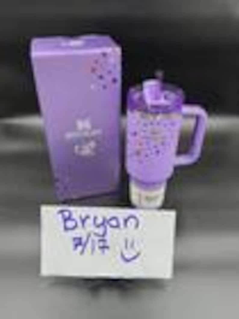 Puede incluir: Un vaso t&eacute;rmico morado con asa, tapa abatible y base blanca, junto a una caja morada a juego. El vaso y la caja presentan un dise&ntilde;o moteado. Una nota manuscrita con el nombre "Bryan" y la fecha 7/17 est&aacute; en la imagen.