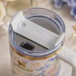 May include: Close-up of a floral-patterned Stanley mug with a white lid and handle. The mug features a gold rim and the Stanley logo. The floral design includes pink, blue, and white hues.