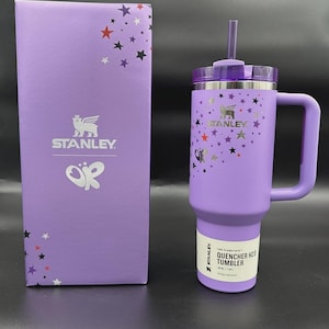 Puede incluir: Un vaso Stanley Quencher H2.0 morado con asa, pajita y tapa, junto a una caja a juego. El vaso y la caja presentan dise&ntilde;os de estrellas y mariposas. El vaso tiene el logotipo de Stanley y el texto "QUENCHER H2.0 TUMBLER".
