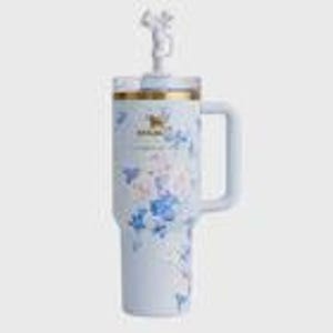 May include: A light blue insulated tumbler with a handle and a decorative lid. The tumbler features a floral pattern in shades of blue, pink, and white. The lid has a small figurine on top. The tumbler has a gold rim and a logo.