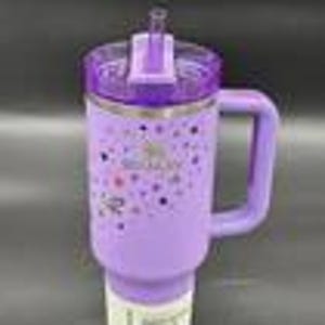 Puede incluir: Un vaso aislado morado con asa y tapa transparente con pajita. El vaso tiene una banda de acero inoxidable y un dise&ntilde;o de estrellas de colores y la palabra "Stanley". El vaso est&aacute; dise&ntilde;ado para caber en un portavasos de coche.