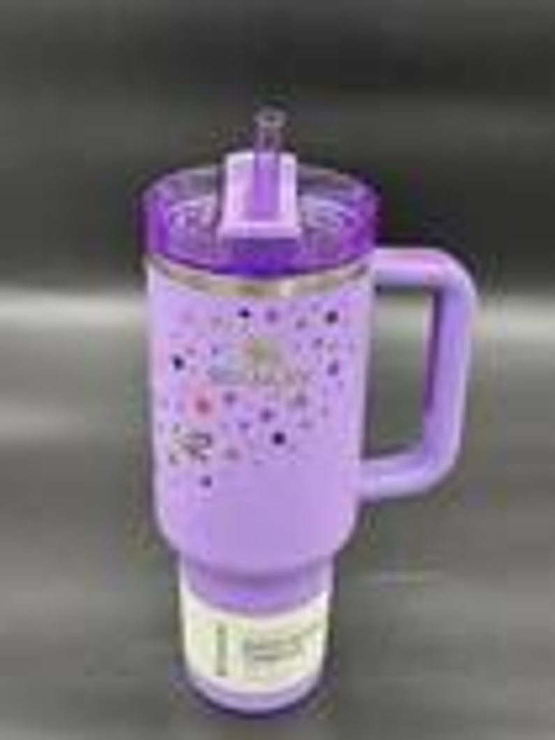 Puede incluir: Un vaso aislado morado con asa y tapa transparente con pajita. El vaso tiene una banda de acero inoxidable y un dise&ntilde;o de estrellas de colores y la palabra "Stanley". El vaso est&aacute; dise&ntilde;ado para caber en un portavasos de coche.