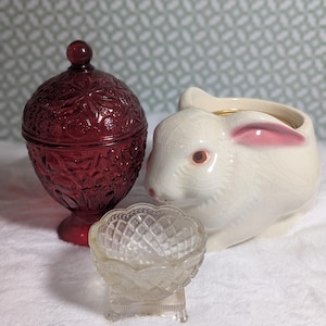 Könnte beinhalten: Eine rubinrote Bonbonniere mit Deckel und floralem Muster, eine kleine, klare Glasschale und ein weißer Keramik-Hasen-Pflanztopf mit rosa Akzenten. Die Objekte stehen auf einer weißen Oberfläche.
