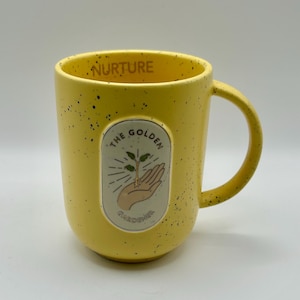 May include: Yellow ceramic mug with black speckles and a curved handle. The mug features an oval graphic with a hand holding a sprouting plant, and the text "THE GOLDEN GARDENER". The word "NURTURE" is printed on the rim.
