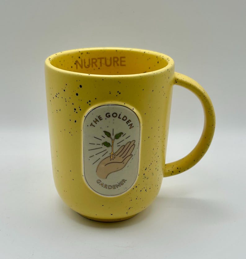 May include: Yellow ceramic mug with black speckles and a curved handle. The mug features an oval graphic with a hand holding a sprouting plant, and the text "THE GOLDEN GARDENER". The word "NURTURE" is printed on the rim.