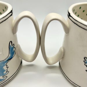May include: Two white ceramic mugs with curved handles. Each mug features a black rim and a blue bird design. The interior of one mug has green polka dots. The mugs are set against a white background.