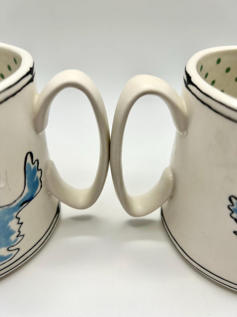 May include: Two white ceramic mugs with curved handles. Each mug features a black rim and a blue bird design. The interior of one mug has green polka dots. The mugs are set against a white background.