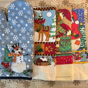 May include: A light blue oven mitt featuring a snowman, snowflakes, and a red bird, alongside three kitchen towels. The towels display a moose in a snowy forest, a figure carrying a green sack, and a snowman with a Christmas tree design.