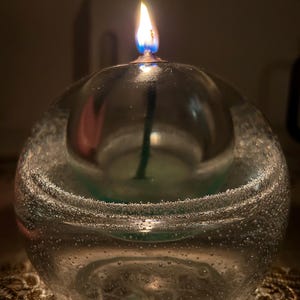 May include: A lit glass candle in a round, bubble-filled container. The flame is yellow and blue, and the candle is set on a decorative surface. The candle is a decorative item.