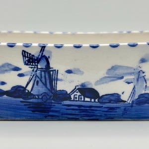 May include: A rectangular ceramic planter with a white base and blue hand-painted illustrations of a windmill, a small house, and clouds. The top rim features a decorative blue dotted pattern, and the base has a solid blue painted border.