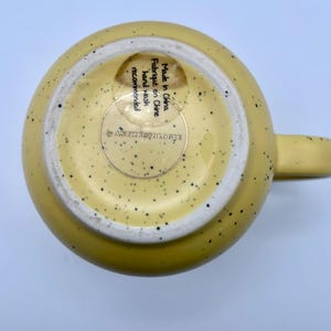 May include: A yellow ceramic mug with a speckled design and a handle. The bottom of the mug has a white interior and a sticker that reads "Made in China, hand wash recommended" and the brand name.