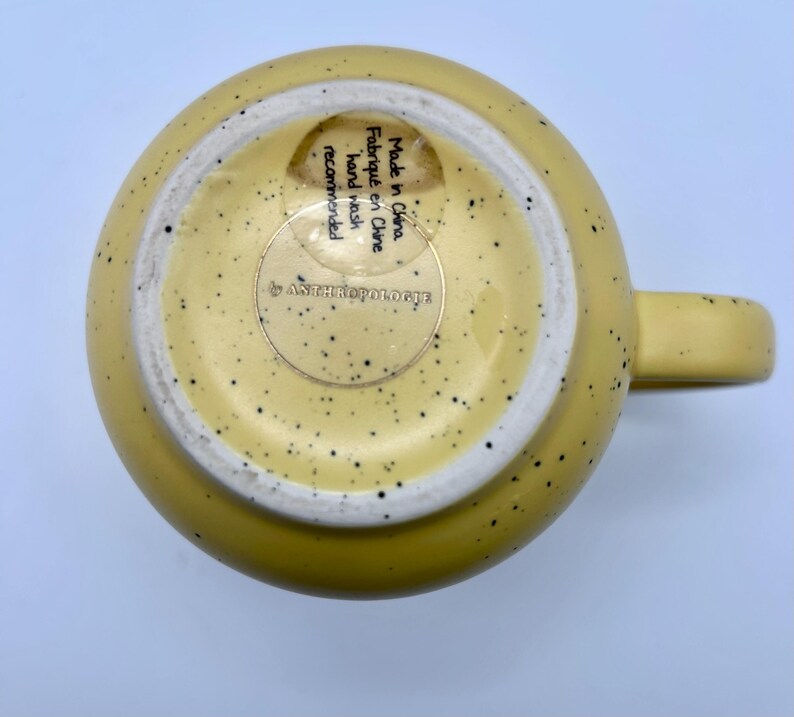 May include: A yellow ceramic mug with a speckled design and a handle. The bottom of the mug has a white interior and a sticker that reads "Made in China, hand wash recommended" and the brand name.