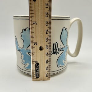 May include: A cream-colored ceramic mug with a blue and black design. The mug is approximately 4 inches tall. The design includes a blue shape and the word "hello" in black script. The mug has a curved handle.