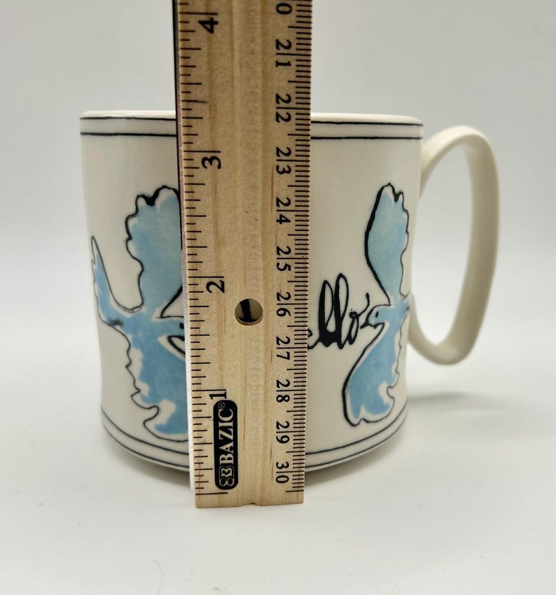 May include: A cream-colored ceramic mug with a blue and black design. The mug is approximately 4 inches tall. The design includes a blue shape and the word "hello" in black script. The mug has a curved handle.