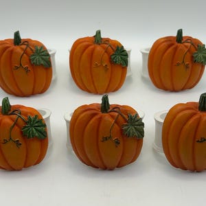 May include: A set of six orange pumpkin napkin rings, each featuring a green stem and a single green leaf. The pumpkins are attached to white circular rings, providing a functional and decorative element for table settings during the autumn season.