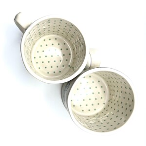 May include: Two ceramic mugs with a cream-colored base and a pattern of small, evenly spaced green dots inside. The mugs have white handles and a slightly flared rim. The mugs are angled towards the viewer.