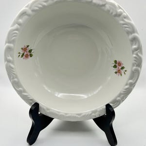 May include: A white ceramic serving bowl with a decorative floral and leaf pattern around the rim. The bowl features small pink floral accents and is displayed on a black stand. A classic piece for serving.