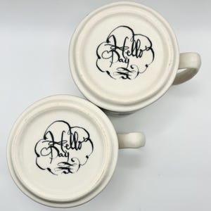 May include: Two white ceramic mugs with handles, bottoms facing up. Each mug has a black, stylized "Hello Day" design inside a cloud-like shape. The mugs are on a white background.