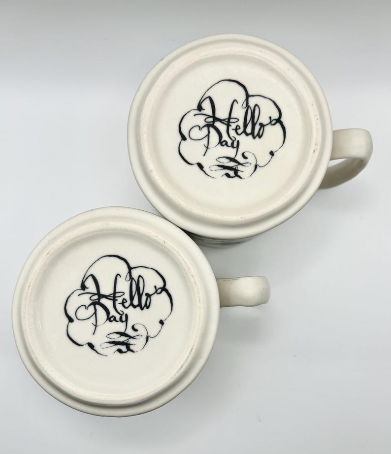 May include: Two white ceramic mugs with handles, bottoms facing up. Each mug has a black, stylized "Hello Day" design inside a cloud-like shape. The mugs are on a white background.