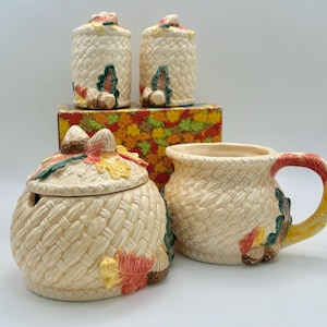 May include: A ceramic kitchen set featuring a lidded sugar bowl, a mug, and two shakers, all with a cream-colored woven basket texture. Each piece is decorated with painted autumn leaves and acorns in shades of red, orange, yellow, and green.