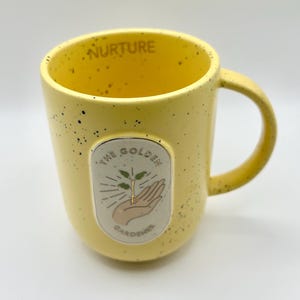 May include: Yellow ceramic mug with black speckles and a curved handle. The mug features an oval emblem with a hand holding a sprouting plant, the words "THE GOLDEN GARDENER" and "NURTURE".