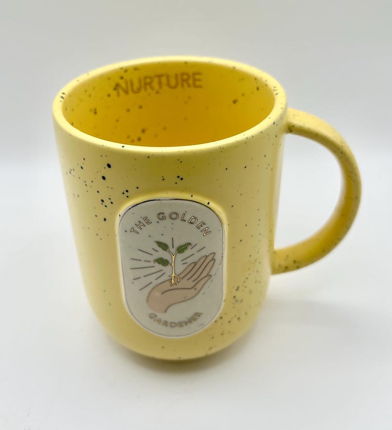 May include: Yellow ceramic mug with black speckles and a curved handle. The mug features an oval emblem with a hand holding a sprouting plant, the words "THE GOLDEN GARDENER" and "NURTURE".