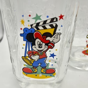 May include: Clear glass featuring a cartoon Mickey Mouse design. Mickey is wearing a red hat, shirt, and shoes, blue pants, and is holding a trumpet. The glass also has a film clapperboard and stars.