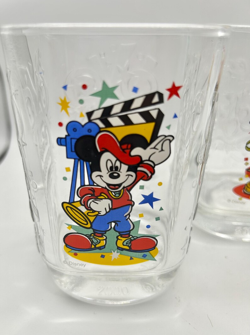 May include: Clear glass featuring a cartoon Mickey Mouse design. Mickey is wearing a red hat, shirt, and shoes, blue pants, and is holding a trumpet. The glass also has a film clapperboard and stars.