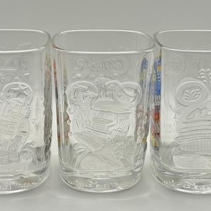 May include: Three clear, square-shaped drinking glasses with embossed designs. Each glass features a different embossed design, including a building, a person, and a decorative pattern. The glasses are displayed on a white surface.