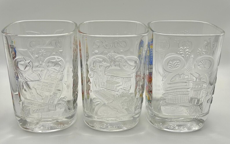 May include: Three clear, square-shaped drinking glasses with embossed designs. Each glass features a different embossed design, including a building, a person, and a decorative pattern. The glasses are displayed on a white surface.