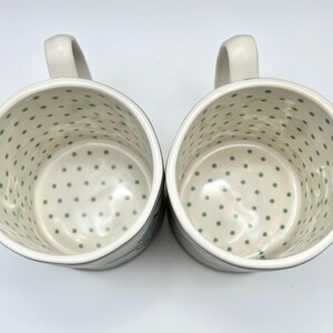 May include: Two white ceramic mugs with a light green polka dot pattern inside. Each mug has a curved handle and a dark rim. The mugs are likely for coffee or tea.