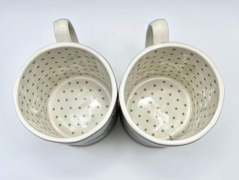 May include: Two white ceramic mugs with a light green polka dot pattern inside. Each mug has a curved handle and a dark rim. The mugs are likely for coffee or tea.