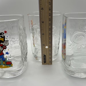 May include: Three clear glass tumblers with embossed designs. One features Mickey Mouse with a film reel, another has a bird design, and the third has a floral pattern. A ruler shows the tumblers are approximately 5 inches tall.