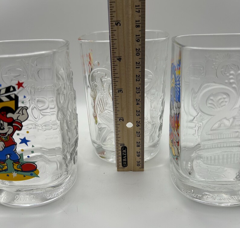 May include: Three clear glass tumblers with embossed designs. One features Mickey Mouse with a film reel, another has a bird design, and the third has a floral pattern. A ruler shows the tumblers are approximately 5 inches tall.