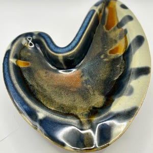May include: A ceramic ashtray with a unique, organic shape. The ashtray features a blend of colors, including blue, brown, orange, and cream. The interior has a textured appearance, and the rim is a darker blue.