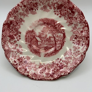 May include: A white and red decorative plate with a scalloped edge. The plate features a detailed red floral pattern around the rim and a central scene of a water mill and trees.