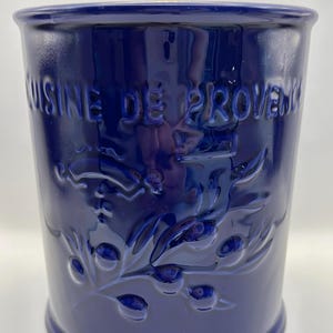 May include: A deep cobalt blue cylindrical ceramic utensil holder with a glossy finish. The exterior features embossed text reading CUISINE DE PROVENCE above a relief design of an olive branch with leaves and small round olives.
