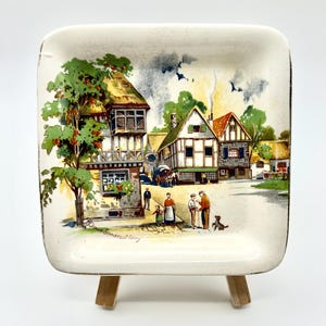 May include: Square ceramic dish with a painted village scene. The artwork depicts buildings with thatched roofs, trees, and figures. The dish is off-white with a slightly aged appearance, and is displayed on a small wooden easel.