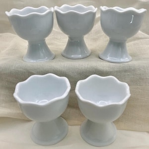 May include: A set of five white ceramic egg cups, each featuring a smooth, glossy finish and a distinct scalloped, wavy rim. The cups are designed with a rounded bowl shape that tapers down into a sturdy, flared pedestal base for stability.