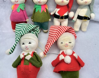 May include: A collection of six felt elf ornaments with painted faces and various colored outfits including red, green, pink, and white. The figures feature pointed hats, some with striped patterns, and attached hanging hooks for display purposes.