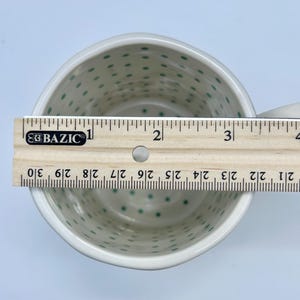 May include: A white ceramic mug with a handle, decorated with small green polka dots. A wooden ruler is placed across the top of the mug, measuring approximately 4 inches in length.