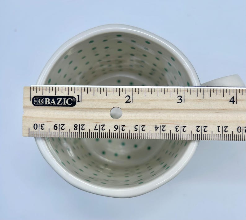 May include: A white ceramic mug with a handle, decorated with small green polka dots. A wooden ruler is placed across the top of the mug, measuring approximately 4 inches in length.