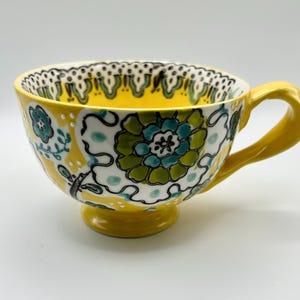 May include: A yellow ceramic teacup featuring a hand-painted floral pattern in shades of teal, lime green, and black outlines. The cup has a wide, flared rim with a black and white dotted border, a pedestal base, and a curved yellow handle.