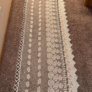 May include: White lace runner with intricate openwork patterns. The design features a combination of geometric shapes, floral motifs, and a scalloped edge. This decorative textile is suitable for various home decor styles.