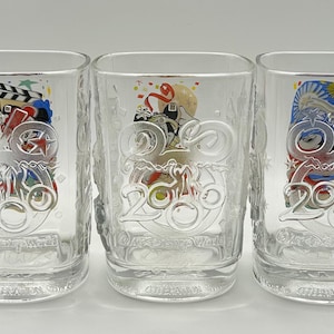 May include: Three clear, square-shaped drinking glasses. Each glass has a decorative design with the number "2006" and colorful images. The glasses are made of clear glass and are suitable for beverages.