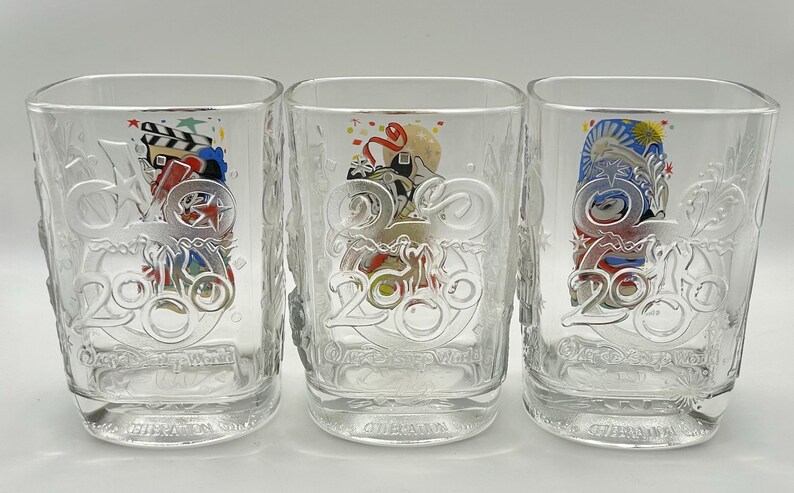 May include: Three clear, square-shaped drinking glasses. Each glass has a decorative design with the number "2006" and colorful images. The glasses are made of clear glass and are suitable for beverages.