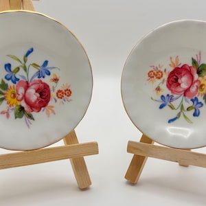 May include: Two small, white porcelain plates with gold rims, each adorned with a floral bouquet of red roses, blue and yellow flowers. The plates are displayed on miniature wooden easels, perfect for decorative purposes.