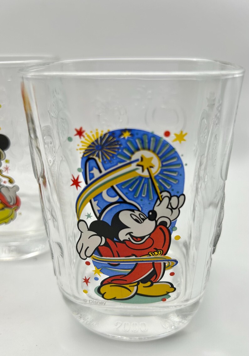 May include: Clear glass tumbler featuring a colorful graphic of Mickey Mouse in a sorcerer's outfit, holding a magic wand. The design includes stars and fireworks. The word "Disney" is printed on the glass.