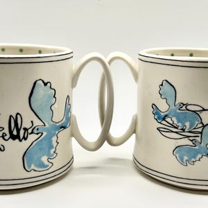 May include: Two white ceramic mugs with a unique handle design, joined together. Each mug features a blue bird design and black text that says "Hello". The rims are decorated with a black line and green dots.